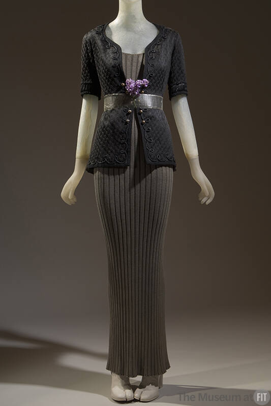 Black elbow length floral brocade jacket over floor length grey ribbed sweater dress and purple…