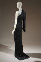 Rear view of black asymmetrical ensemble showing lapels on right side of jacket and skirt train