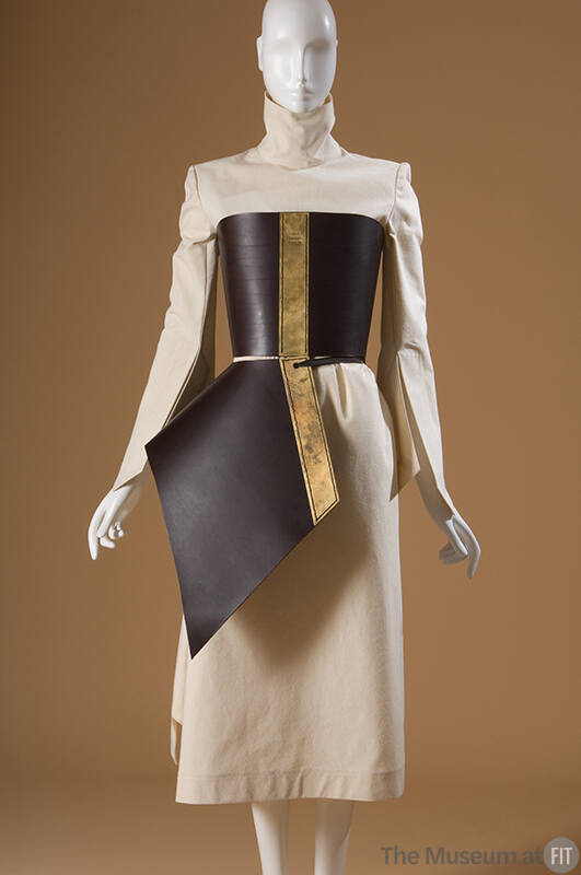 The Aketon beige dress with high neck and flared, pointed sleeves; brown corset and apron with …