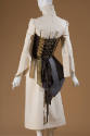 Back of the Aketon beige dress with high neck and flared, pointed sleeves; brown corset and apr…