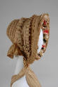 Side view of beige bonnet trimmed with lace and cloth flowers inside brim, neck tie