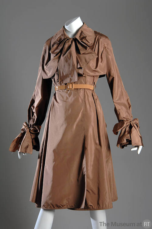 Taupe knee length rain coat with large bow at neck and long sleeve cuffs, tan leather belt
