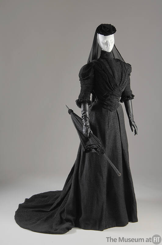 Black crinkle fabric jacket with high neck, floor length skirt with train, hat with veil and ma…