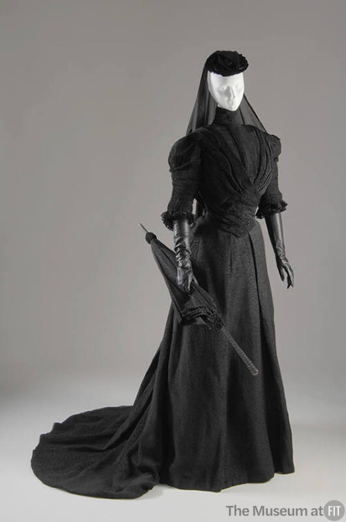 Black crinkle fabric jacket with high neck, floor length skirt with train, hat with veil and ma…