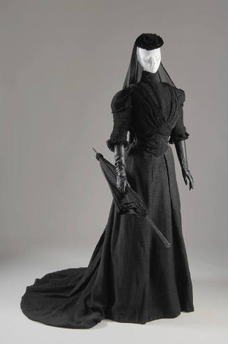 Black crinkle fabric jacket with high neck, floor length skirt with train, hat with veil and ma…