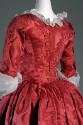 Detail view of back of red damask dress showing white neck and sleeve ruffles, seams from shoul…