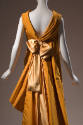 Back view of orange evening gown showing deep V-back with oversize pale orange bow at center an…