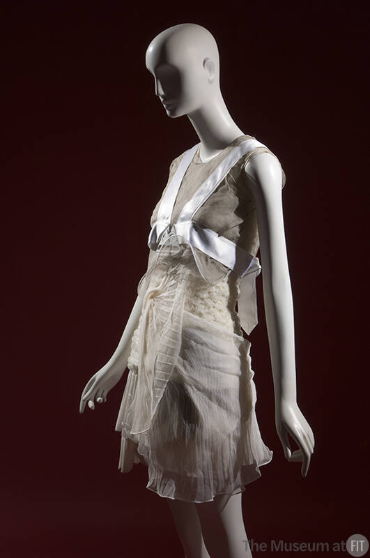 ¾ view of ensemble with grey sheer sleeveless cropped top, white pleated mini dress and white h…