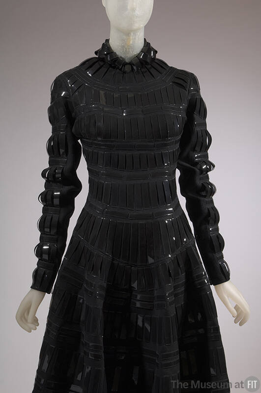 Detail front view of black wool dress with black patent leather tabs appliqued in grid pattern,…