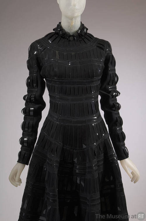 Detail front view of black wool dress with black patent leather tabs appliqued in grid pattern,…