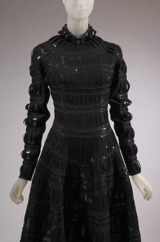 Detail front view of black wool dress with black patent leather tabs appliqued in grid pattern,…