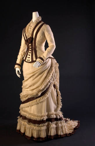 ¾ view of two-piece ivory dress with brown princess seam trim on long sleeve bodice and brown t…