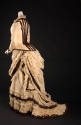 Back view of two-piece ivory dress showing brown trim down center of bodice and draped bustle a…