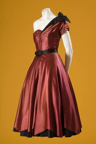 Red satin evening dress with asymmetrical neckline, black belt at waist, and ribbon bow on left…
