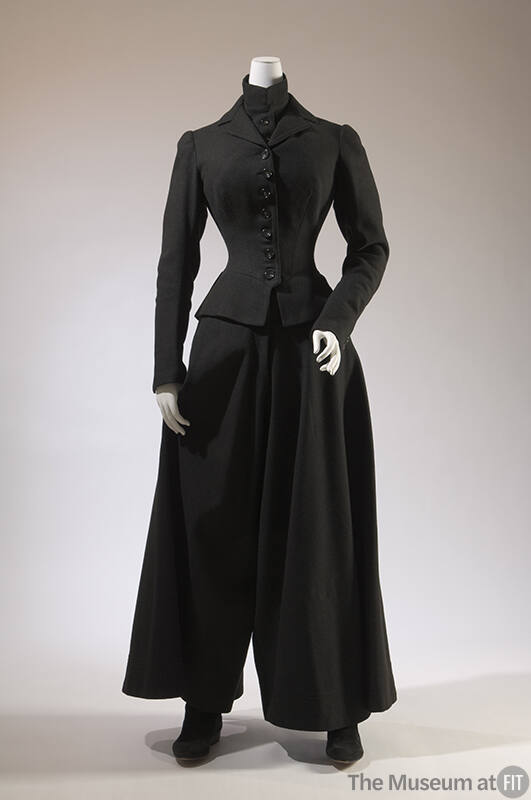 Black ensemble of fitted button front jacket over band collar chest warmer with wide flared leg…