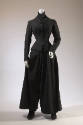 Black ensemble of fitted button front jacket over band collar chest warmer with wide flared leg…