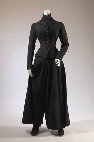 Black ensemble of fitted button front jacket over band collar chest warmer with wide flared leg…