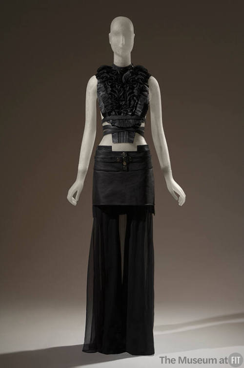 Black leather pleated top with black ruffled bib front and crisscross leather straps at waist, …