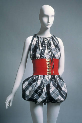 Sleeveless black and white plaid romper with puffed legs, dressed with wide red cinch belt with…