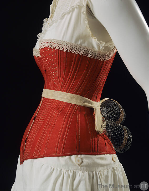 Detail view of red corset shown with white laced trimmed combination petticoat and bustle of do…