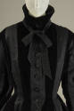 Close up of the bodice of a black coat with a high collar with bow, buttons down center-front, …