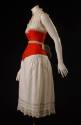 Red corset shown with white laced trimmed combination petticoat and bustle of double crescent t…
