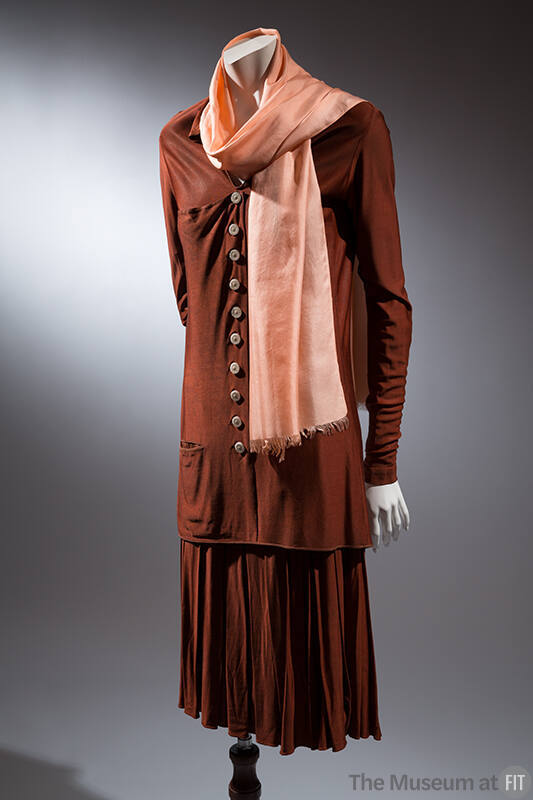 Brown skirt suit with tunic-length buttoned blouse with long sleeves, loosely pleated skirt and…