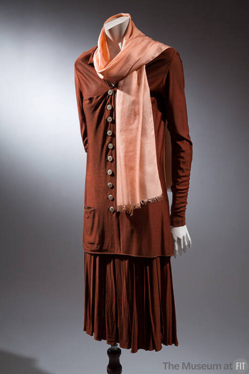 Brown skirt suit with tunic-length buttoned blouse with long sleeves, loosely pleated skirt and…