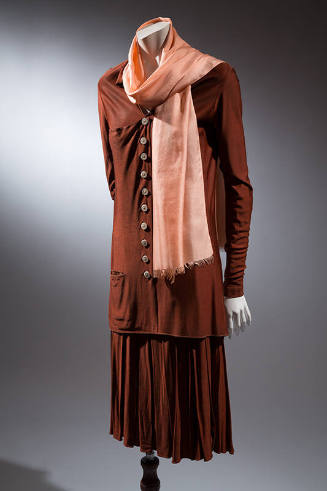 Brown skirt suit with tunic-length buttoned blouse with long sleeves, loosely pleated skirt and…