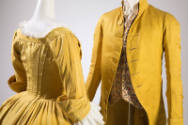 Detail view of yellow/green dress with fitted bodice and bustle skirt on right and man’s cutawa…