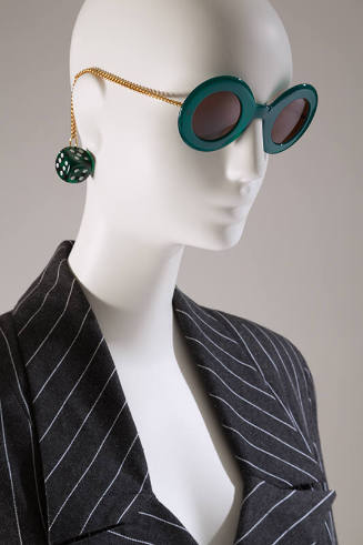 Green sunglasses with round frame and gold metal chain arms ending in green dice with grey and …