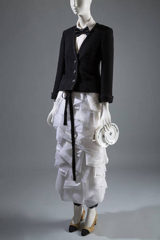 Ensemble of black jacket, white shirt, black bow tie, white ruffled ankle length skirt, beige p…