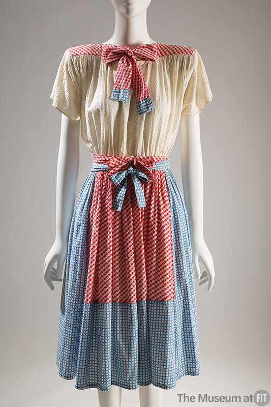 Knee-length dress with off-white blouson bodice and blue gingham skirt and tie with red gingham…
