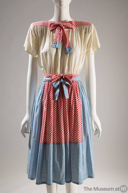 Knee-length dress with off-white blouson bodice and blue gingham skirt and tie with red gingham…