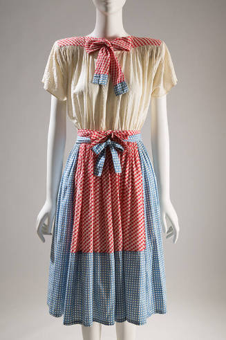 Knee-length dress with off-white blouson bodice and blue gingham skirt and tie with red gingham…