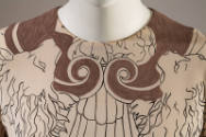 Detail view of taupe dress neckline showing taupe scroll pattern and white and black outlined l…