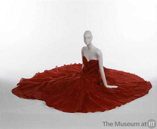 Seated mannequin in red strapless dress with petal-like layers, full skirt spread out
