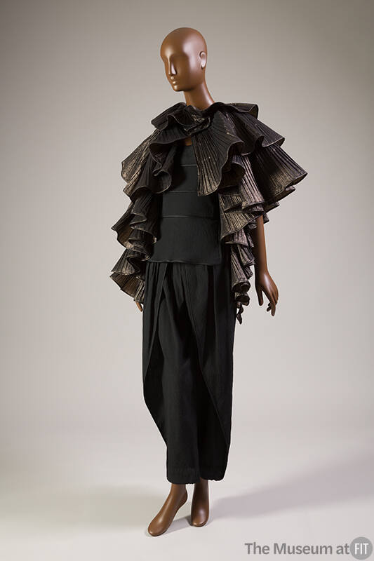 Black metallic pleated cape in ruffled tiers over black tiered peplum top and matching tapered …