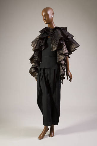 Black metallic pleated cape in ruffled tiers over black tiered peplum top and matching tapered …
