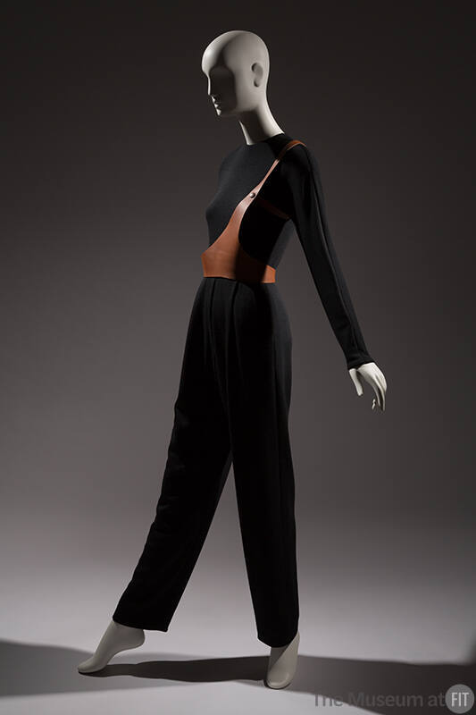 Black long sleeve jumpsuit with one strap brown leather harness over right shoulder and around …