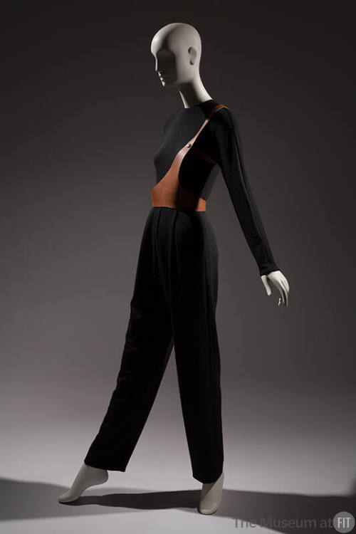 Black long sleeve jumpsuit with one strap brown leather harness over right shoulder and around …