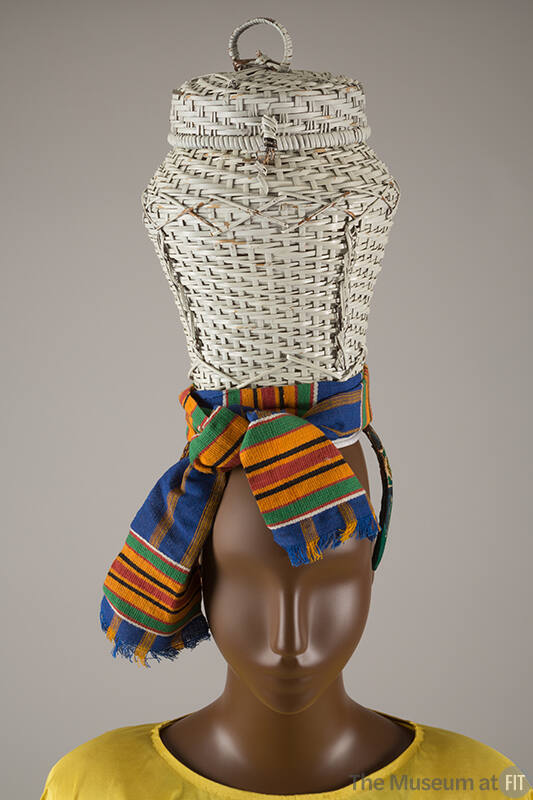 Detail view of wicker basket hat showing green, orange, black, red and white Kente cloth sash t…