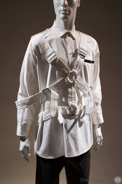 Man’s white oxford style shirt with multiple buckled straps overtop and linked with two front r…