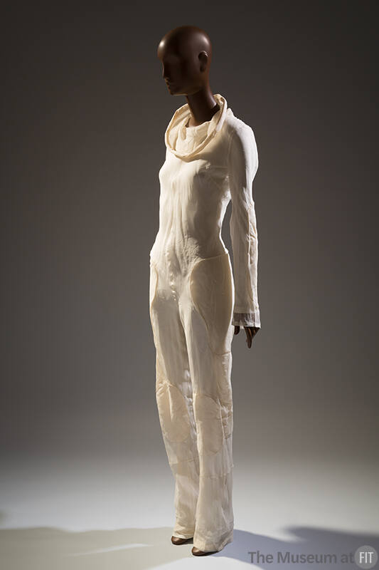 Ivory jumpsuit with funnel cowl neckline, long sleeves and padded patches at hip, thigh and kne…