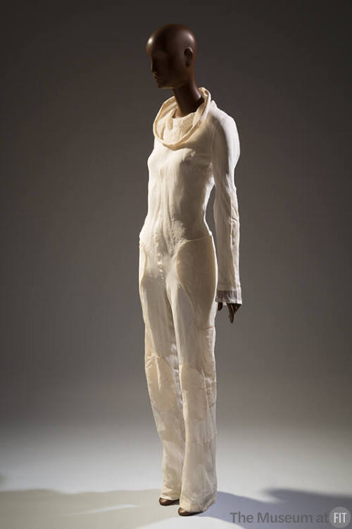 Ivory jumpsuit with funnel cowl neckline, long sleeves and padded patches at hip, thigh and kne…