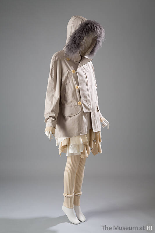 Beige coat with button and loop closure and hood trimmed in grey fur over off-white dress with …