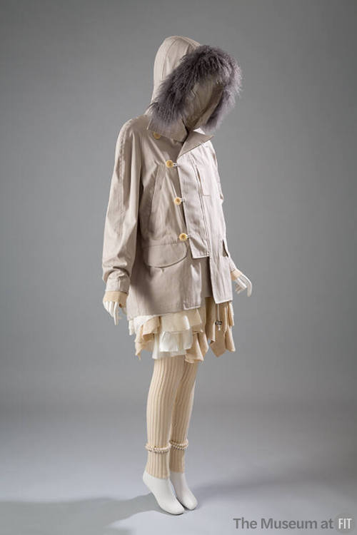 Beige coat with button and loop closure and hood trimmed in grey fur over off-white dress with …