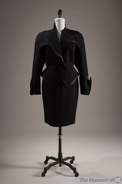 Black suit of fitted jacket with peplum, asymmetrical collar and cuffs and faux leather trim an…