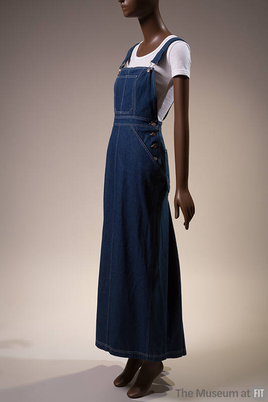 Dark blue denim floor length overalls dress with white topstitching over white short sleeve t-s…