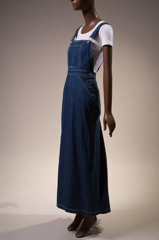 Dark blue denim floor length overalls dress with white topstitching over white short sleeve t-s…